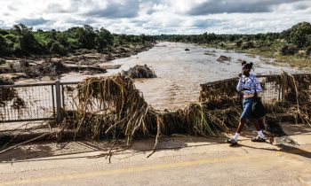 Flooded river blocking a road with a woman walking past debris.